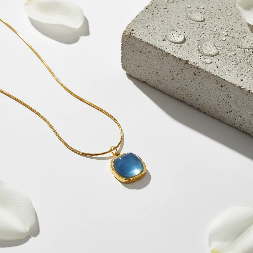 A close-up, high-fashion product shot of a gold necklace with a square blue gemstone pendant, against a minimalist