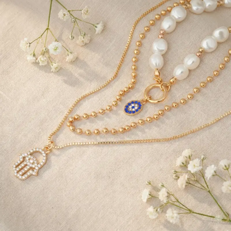 Gold necklace with a hamsa hand charm and blue evil eye pendant on a beige fabric background with white flowers.