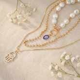 Gold necklace with a hamsa hand charm and blue evil eye pendant on a beige fabric background with white flowers.