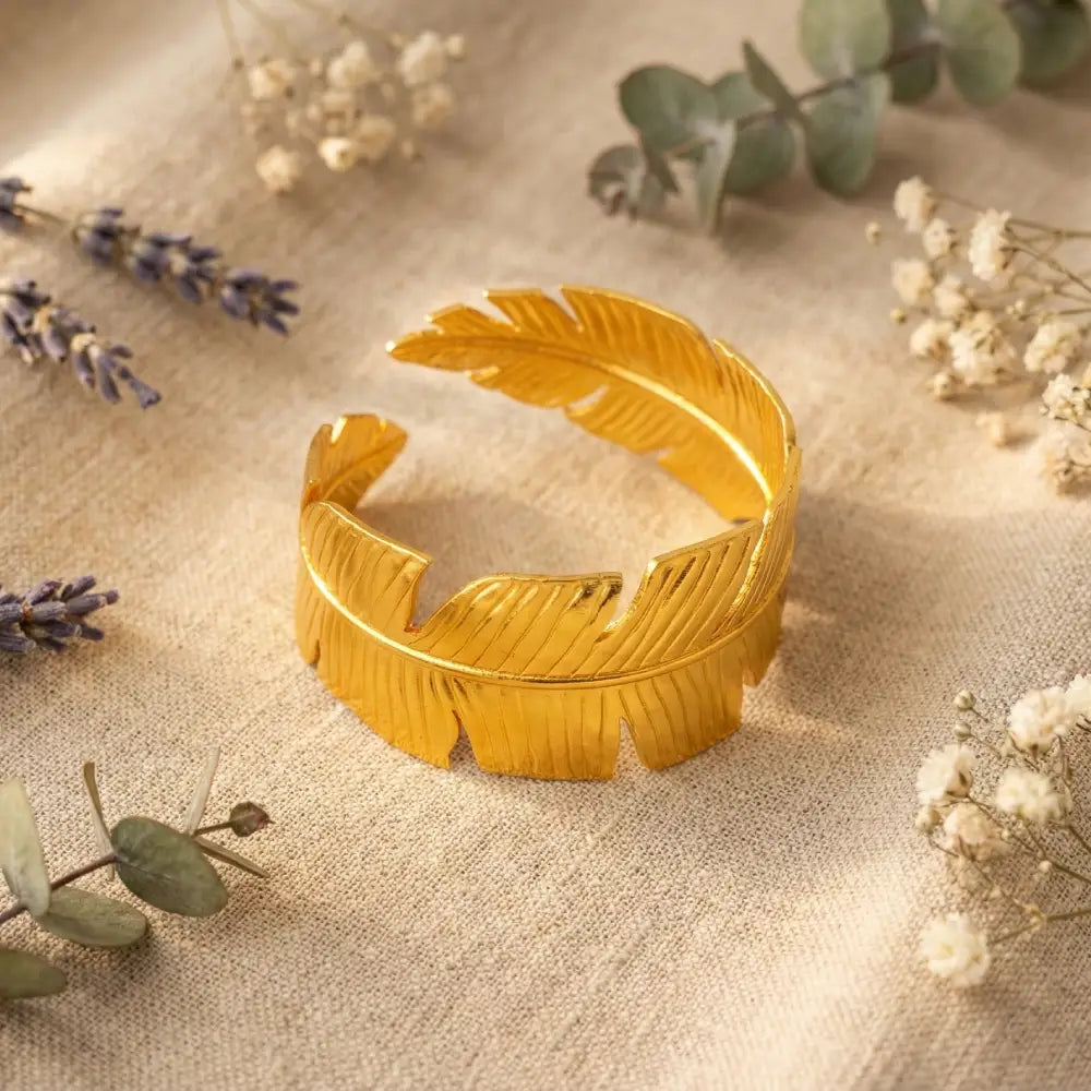 Gold feather-shaped bracelet on a textured surface with dried flowers.