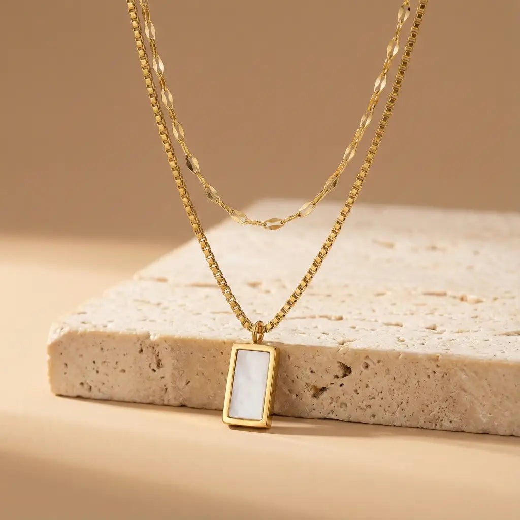 Gold necklace with a rectangular pendant on a beige stone surface